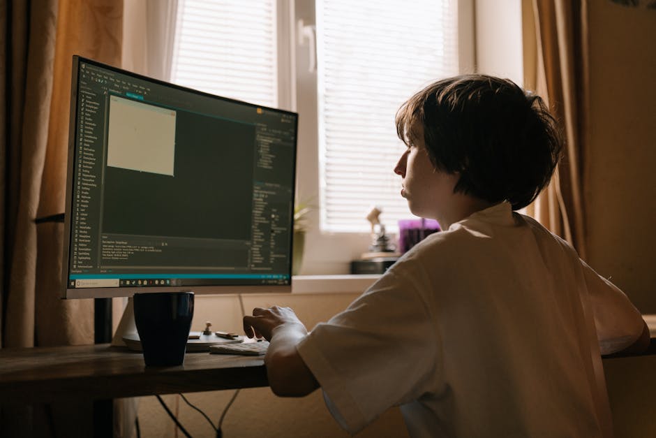 A person with short dark hair, wearing a white shirt, sitting at a wooden desk in front of a large computer monitor, working on gardening or landscaping project planning. The screen displays a dark-th